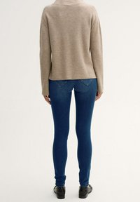 Suéter de punto beige con un ajuste holgado, combinado con jeans ajustados oscuros y zapatos negros, visto desde atrás.