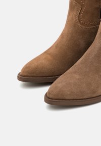 Botine de piele suflată, de culoare brown, cu vârfuri ascuțite, cusături contrastante și o talpă plată de culoare brown. Materialul prezintă o suprafață moale, texturată.