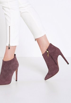 Bottines en daim violettes avec un bout pointu, un talon aiguille et un détail de fermeture éclair sur le côté, assorties à un pantalon blanc zippé.