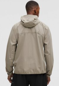 Hellbeige wasserdichte Jacke mit Kapuze, die eine glatte Textur und minimalistisches Design aufweist. Sichtbare Nähte und ein Logo-Akzent nahe dem Saum.