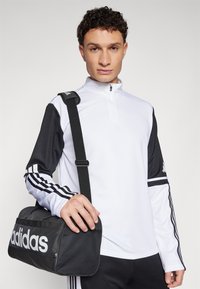 Hvid halv-zip sportstop med sorte ærmer og tre-stribe detaljer. Modellen holder en sort adidas duffeltaske med et stort hvidt logo.