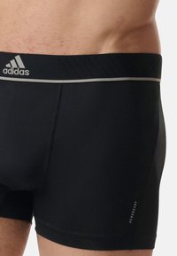 Črne atletske bokserice iz gladkega materiala, z kontrastnim sivim pasom z logotipom "adidas" in napisom "AEROREADY" na strani.