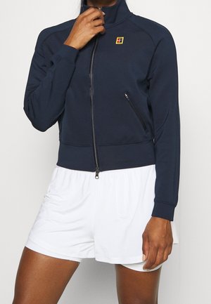 Persona vistiendo chaqueta azul marino con cremallera y logotipo en el pecho, y shorts deportivos blancos, sosteniendo la cremallera de la chaqueta cerca del mentón.