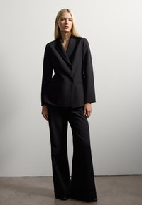 Blazer noir ajusté avec un décolleté en V profond et des épaules structurées, associé à un pantalon noir taille haute et jambes larges, le tout en tissu lisse.