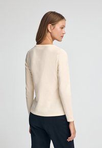 Langärmliges beiges Oberteil aus weichem Material, mit rundem Ausschnitt und taillierter Silhouette, getragen mit dunklen Hosen.