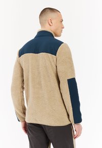 Muž otočený zády, oblečený v béžovém fleecovém kabátu s tmavě modrými ramenními a loketními záplatami, doplněný dvoubarevnými hnědými kalhotami.