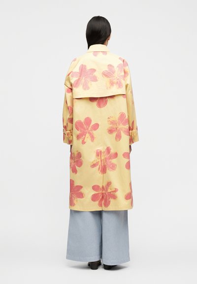 Γυναίκα που φοράει κίτρινο καμπαρντίνα με μεγάλα ροζ λουλούδια, συνδυασμένο με φαρδιά ανοιχτό μπλε παντελόνια και μαύρα παπούτσια, στέκεται με την πλάτη σε εμάς.