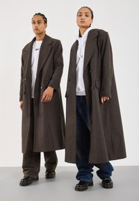 Brown double-breasted long coat in a wool-blend fabric, featuring large lapels, side pockets, and button detailing on the cuffs.