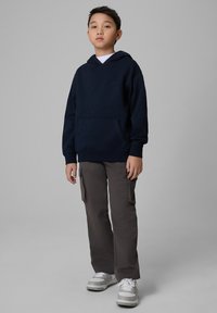 Garçon debout portant un sweat à capuche bleu marine, un pantalon cargo gris et des baskets blanches sur un fond gris uni.