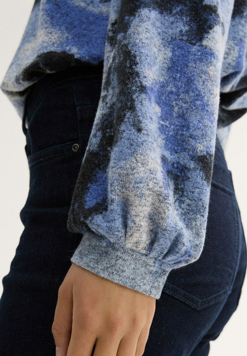 Blau- und graumusterter Strickpullover mit Ballonärmeln und gerippten Bündchen, kombiniert mit dunkelblauen Jeans im Denim-Look mit einer Rückentaschendetail.