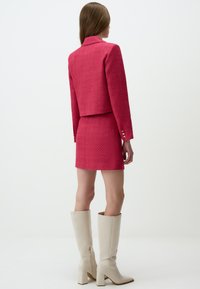Costume rouge texturé avec une veste courte et une jupe assortie, associé à des bottes beige au genou avec un talon bloc et une fermeture éclair sur le côté.
