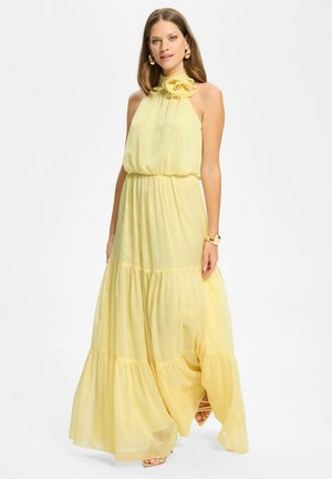 Kobieta ubrana w żółtą, długa do ziemi suknię bez rękawów z wysokim, falbankowym kołnierzem i warstwową spódnicą, stojąca na białym tle.