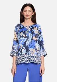 Blouse à motifs floraux bleus avec manches 3/4, présentant un col froncé et un ourlet contrastant à motifs. Associée à un pantalon bleu.