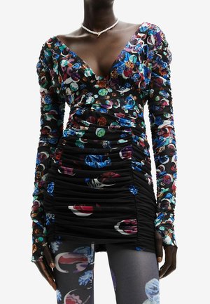 Kobieta ubrana w marszczoną czarną mini sukienkę oraz bluzkę z długim rękawem w kolorowe abstrakcyjne kwiatowe wzory oraz naszyjnik z pereł.