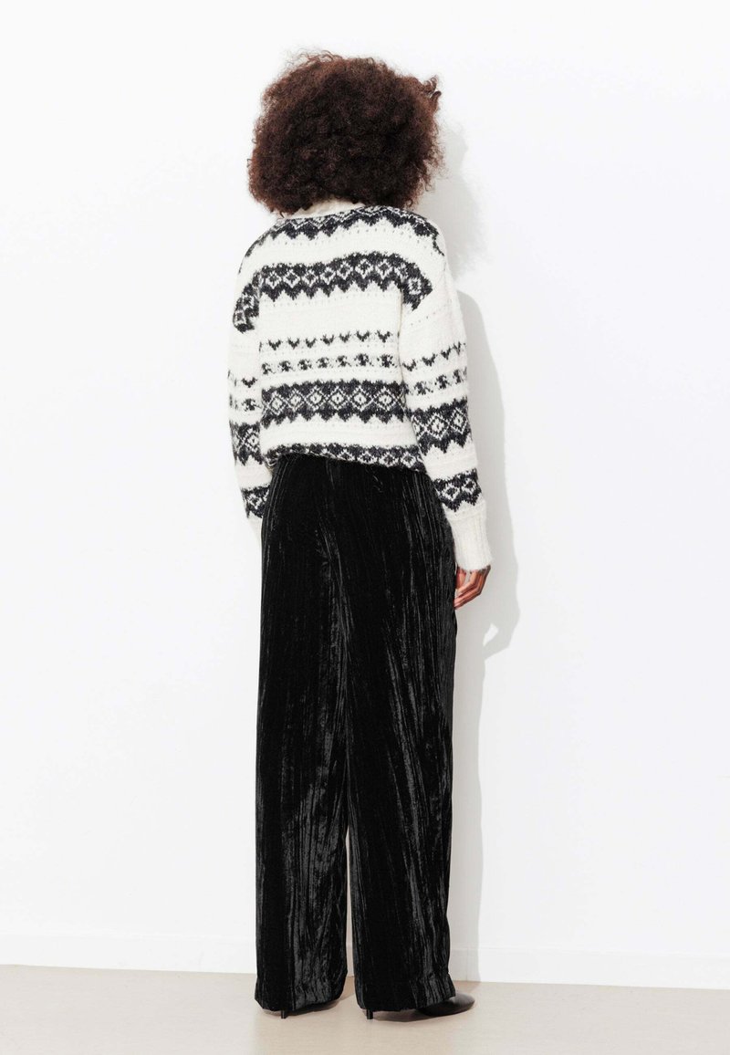 Pull en tricot blanc avec motifs géométriques noirs, associé à un pantalon large en velours noir. Texture douce, coupe décontractée.