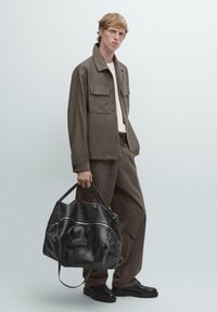 Giacca marrone in misto lana con due tasche frontali, abbinata a pantaloni coordinati. Il modello tiene una borsa da viaggio in pelle nera con cerniere e tracolle.