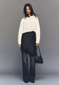 Bijeli skraćeni džemper, crna višeslojna haljina s čipkastim rubom, tamne široke traperice, držeći crnu zakrivljenu torbu. Sivi zid u pozadini.