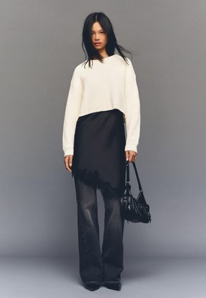 Pulover alb scurt, rochie neagră în straturi cu margine din dantelă, pantaloni largi din denim închis, ținând o geantă neagră curbată. Fundal gri.