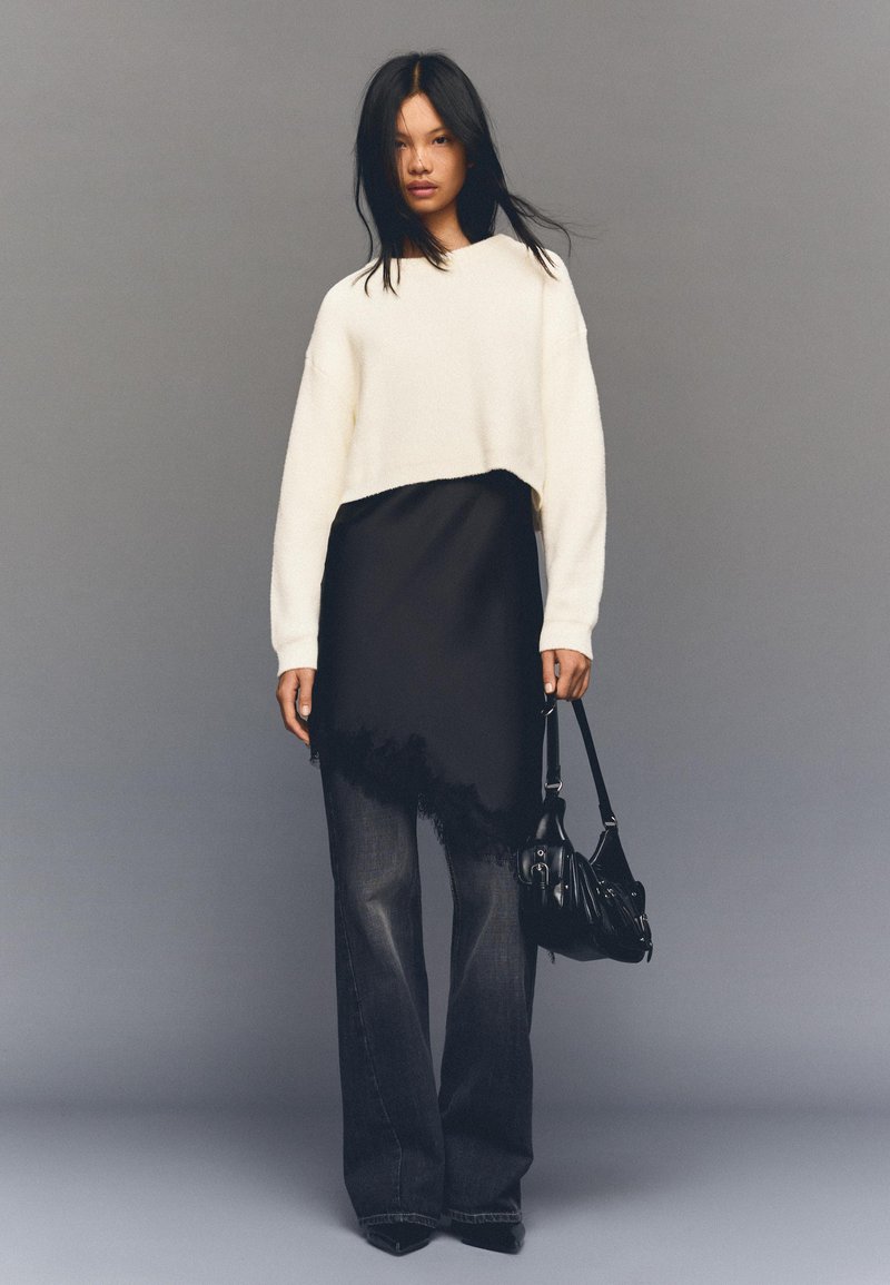 Bijeli skraćeni džemper, crna višeslojna haljina s čipkastim rubom, tamne široke traperice, držeći crnu zakrivljenu torbu. Sivi zid u pozadini.