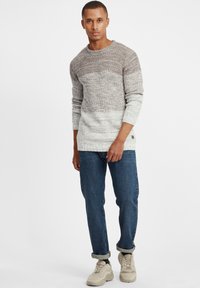 Lichtgrijze en beige gebreide trui met lange mouwen, ronde halslijn en een getextureerd patroon, gecombineerd met blauwe jeans en beige sneakers.