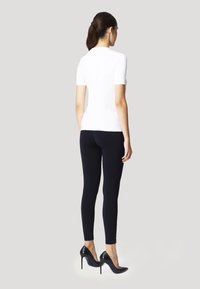 Top bianco a maniche corte abbinato a leggings neri e scarpe nere con tacco alto, mostrato da dietro su uno sfondo grigio chiaro.