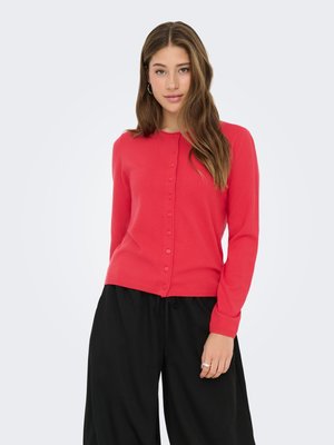 Kardigan na guziki w jaskrawym koralowym kolorze, wykonany z miękkiego materiału. Posiada długie rękawy, okrągły dekolt i pasujące guziki. Noszony z czarnymi spodniami.