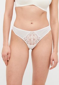 String en dentelle blanche avec un motif floral à l'avant, en tissu transparent, et une taille lisse pour un design minimaliste.