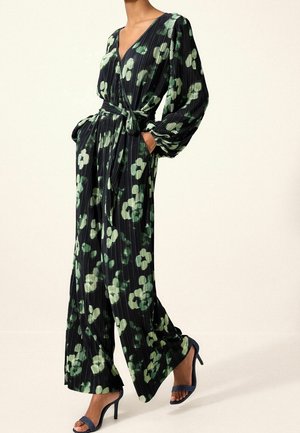 Combinaison plissée noire à motif floral vert, col en V profond, manches longues et taille nouée. Associée à des sandales à talons hauts bleu marine.