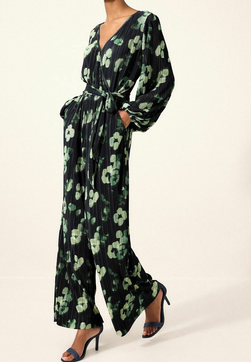 Combinaison plissée noire à motif floral vert, col en V profond, manches longues et taille nouée. Associée à des sandales à talons hauts bleu marine.
