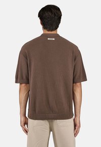 Brauner Pullover mit kurzen Ärmeln, strukturiertem Strick, geripptem Saum und einem kleinen Etikett am Nacken hinten. Das Model trägt helle Hosen.