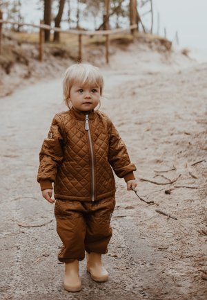 Lastepruun tikandiga jope sobivate pükste ja beežide kummist kummikute komplektis, tolmukivil rajal. Laps hoiab kätt pulgal ja tal on hele blondid juuksed.