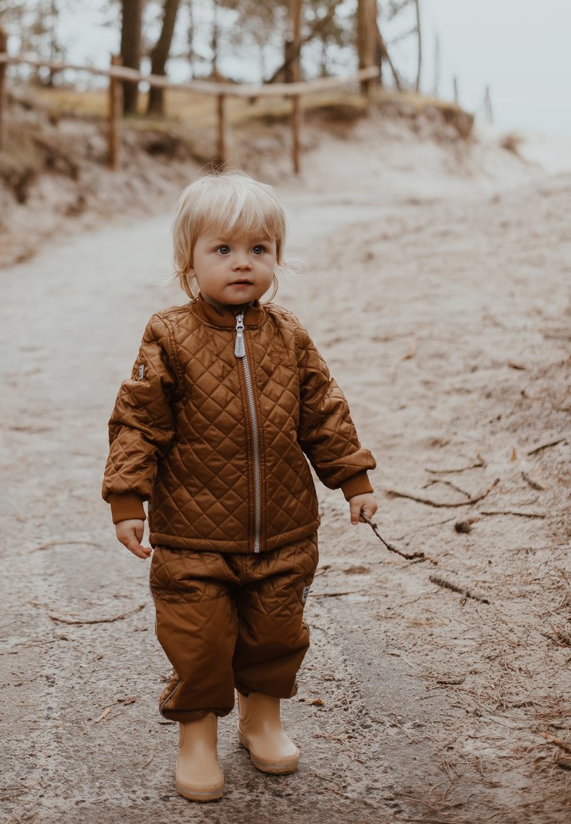 Gyermek barna steppelt dzsekihez illő nadrággal és bézs gumicsizmával, egy földúton. A gyermek botot tart, és világos szőke haja van.