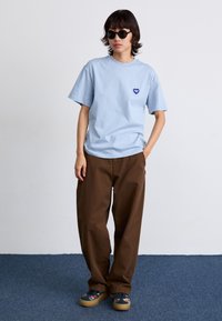 Svijetloplava pamučna majica s logotipom srca, uparen s labavim smeškim hlačama i cipelama s uzorkom. Naočale za sunce upotpunjuju izgled.