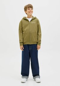 Chłopiec uśmiechający się i stojący, ubrany w oliwkowo-zielony rozpinany hoodie, ciemne szerokie dżinsy z mankietami, białe trampki i białą koszulkę.