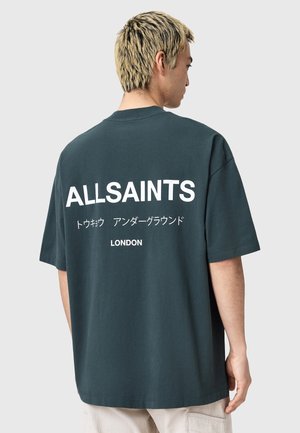 Jeune homme aux cheveux blonds de profil, portant un T-shirt oversized noir avec "ALLSAINTS" et du texte japonais dans le dos.