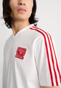 Camiseta de algodão branca com um patch do logótipo da Adidas bordado em vermelho e três riscas verticais vermelhas nos ombros. Design em V.