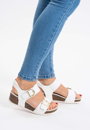 Weiße Keilsandalen mit zwei verstellbaren Riemen, goldenen Schnallen und einer Korkplattform. Die Sohle hat ein strukturiertes Muster für besseren Halt.