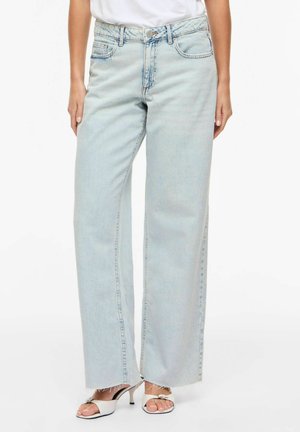 Oseba nosi svetlo modre jeans hlače širokih nogavic z neobdelanimi robovi, v kombinaciji z belimi odprtimi čeveljci in belim zgornjim delom, zataknjenim noter, stoji pred belim ozadjem.