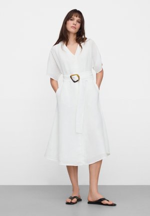 Donna che indossa un vestito midi bianco con scollo a V, maniche corte, cintura con fibbia grande e sandali neri bassi.