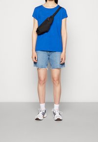 Modrá bavlněná tričko, džínové šortky s třepeným lemem, černá taška přes rameno a bílé tenisky s vícero barvami na šedém pozadí.