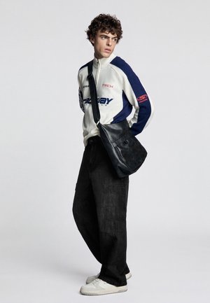 Giovane uomo che indossa un maglione con zip bianco e blu, pantaloni larghi neri, sneakers bianche e una borsa a tracolla nera, guarda di lato.