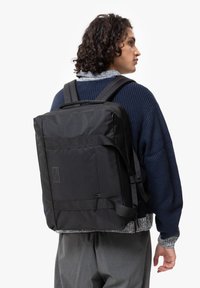 Sac à dos noir de forme rectangulaire, fabriqué en tissu texturé. Comprend des bretelles ajustables et une poche avant avec un patch logo.