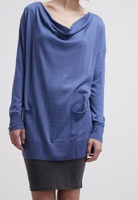 Haut bleu à manches longues avec un col drapé, deux poches frontales et une coupe légèrement ample, associé à une jupe gris foncé.