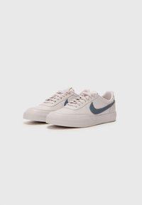 Adidași roz deschis cu o textură netedă, având un swoosh albastru închis, vârf rotund și șireturi plate. Talpa din cauciuc texturat și design minimalist.