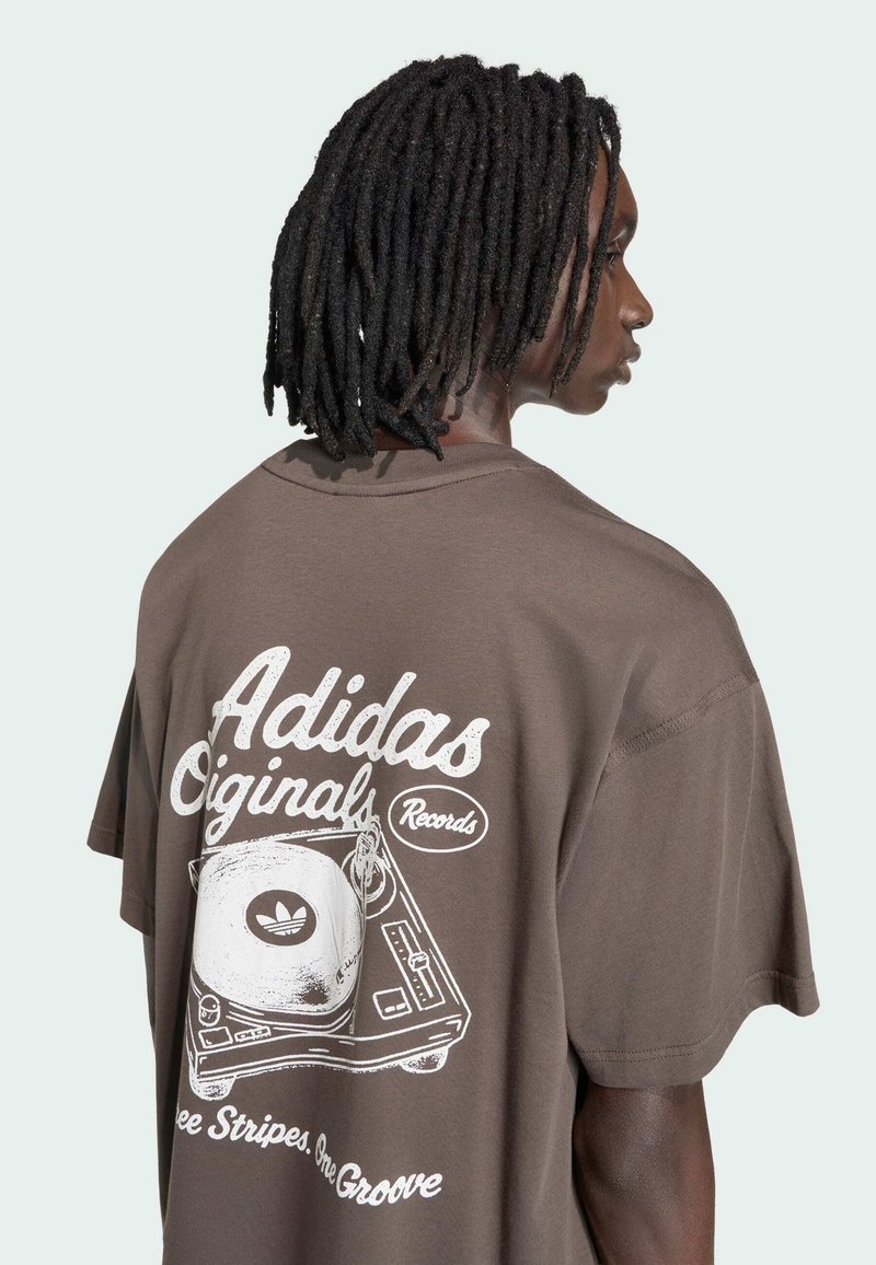 Maglietta oversize marrone con grafica bianca di un giradischi e testo "Adidas Originals Records" sul retro. Maniche corte, materiale in cotone.