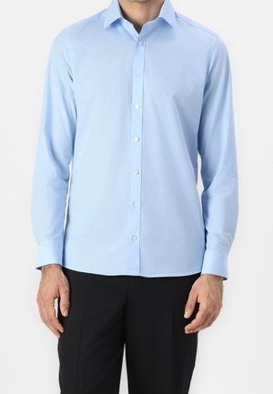 Homme portant une chemise bleu clair à manches longues boutonnée et un pantalon noir sur un fond uni.