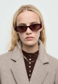 Lunettes de soleil ovales brunes avec des verres foncés, portées par un modèle ayant des cheveux ondulés clairs, associées à un manteau beige par-dessus un haut brun à boutons.