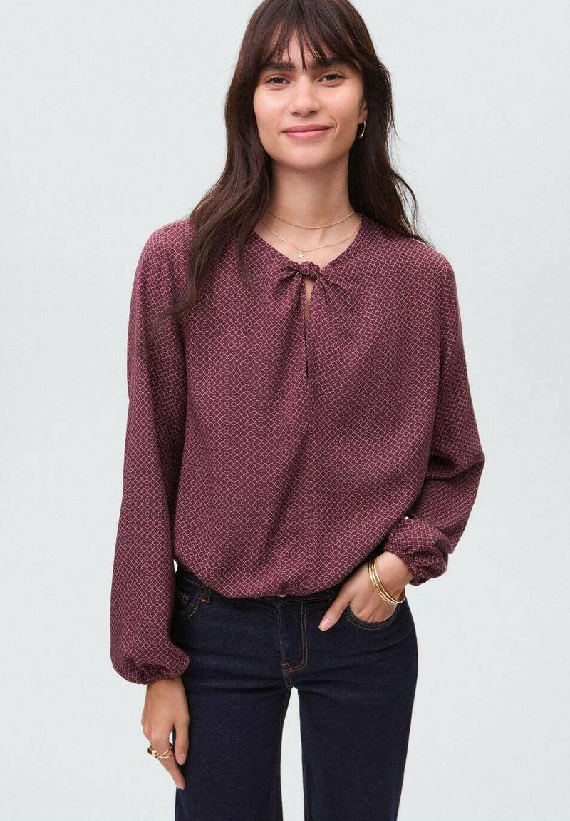Bordeauxfarbene Bluse mit langen Ärmeln, strukturiertem Muster und einem Schlüsselloch-Ausschnitt mit Bindeband, kombiniert mit dunklen Jeans.
