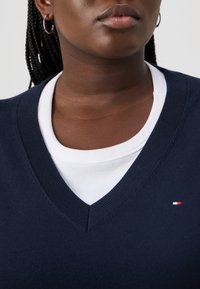 Maglione blu scuro a scollo a V sovrapposto a una maglia bianca a coste, con un piccolo logo rosso, bianco e blu sul lato sinistro. Tessuto morbido al tatto.