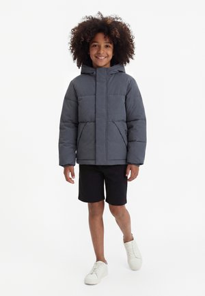 Kind mit lockigem Haar, trägt eine graue Steppjacke mit Kapuze, schwarze Shorts und weiße Turnschuhe, steht und lächelt vor weißem Hintergrund.